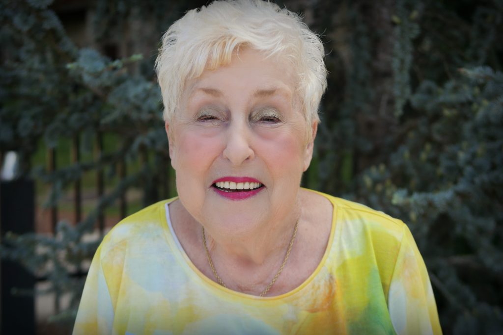 Norma Draper, smiling, looking fabulous with bright pink lipstick and a yellow shirt.